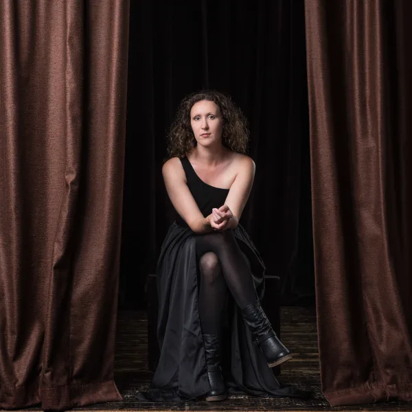 Maegan in a black dress, seated cross-legged in front of a partially-open curtain